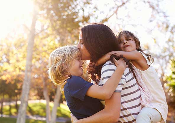 A young mom holds a kid on her back and one on in front of her as they hug her